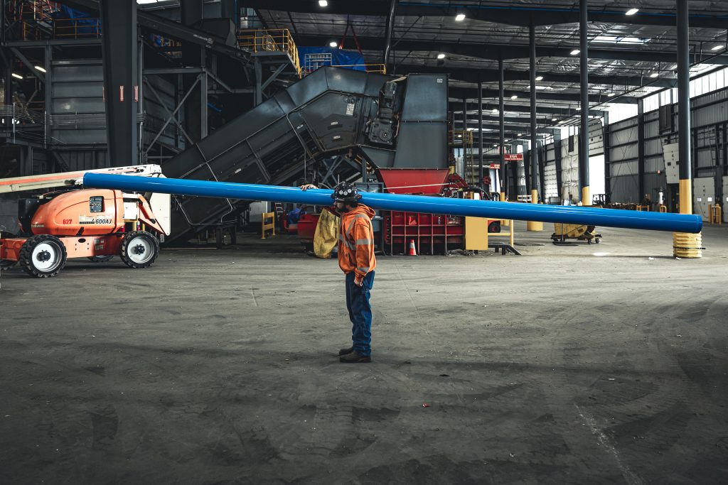 Worker in orange safety vest and helmet holds a long blue pipe in a large industrial facility to the left.