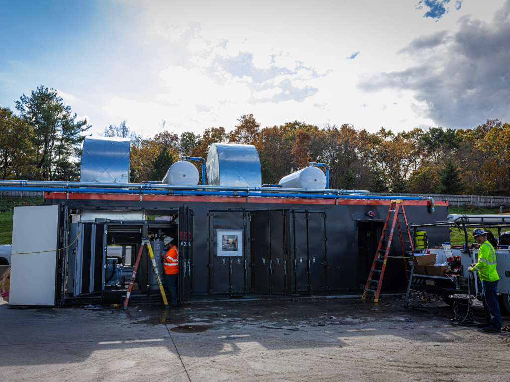 Industrial mobile unit with metal doors and blue piping on top; two workers in safety vests and hard hats nearby, a ladder leaning against the side.