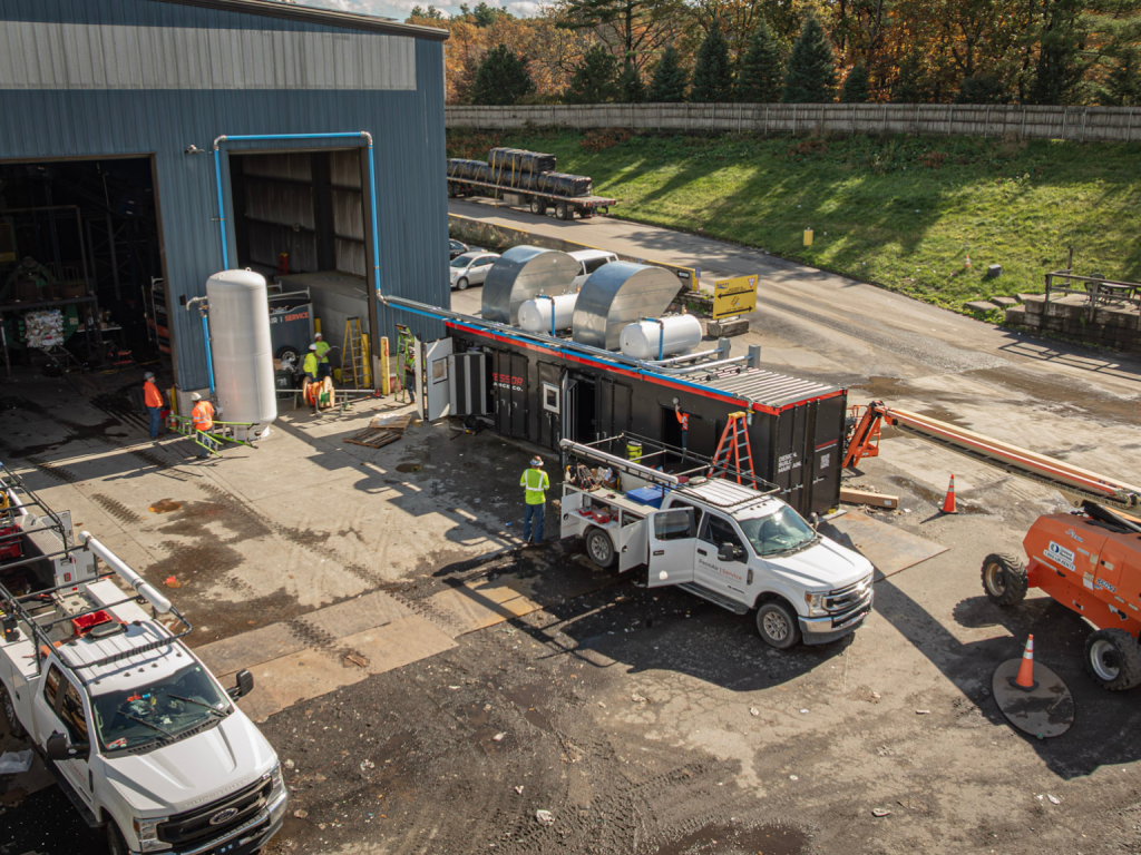 Industrial worksite with a blue metal building, workers in safety vests, and a black modular unit connected to a white service truck outside.