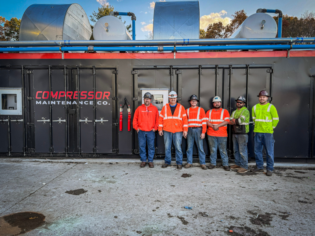 Group of seven maintenance workers in neon safety vests and hard hats standing in front of a large black industrial compressor unit labeled 'COMPRESSOR MAINTENANCE CO.'