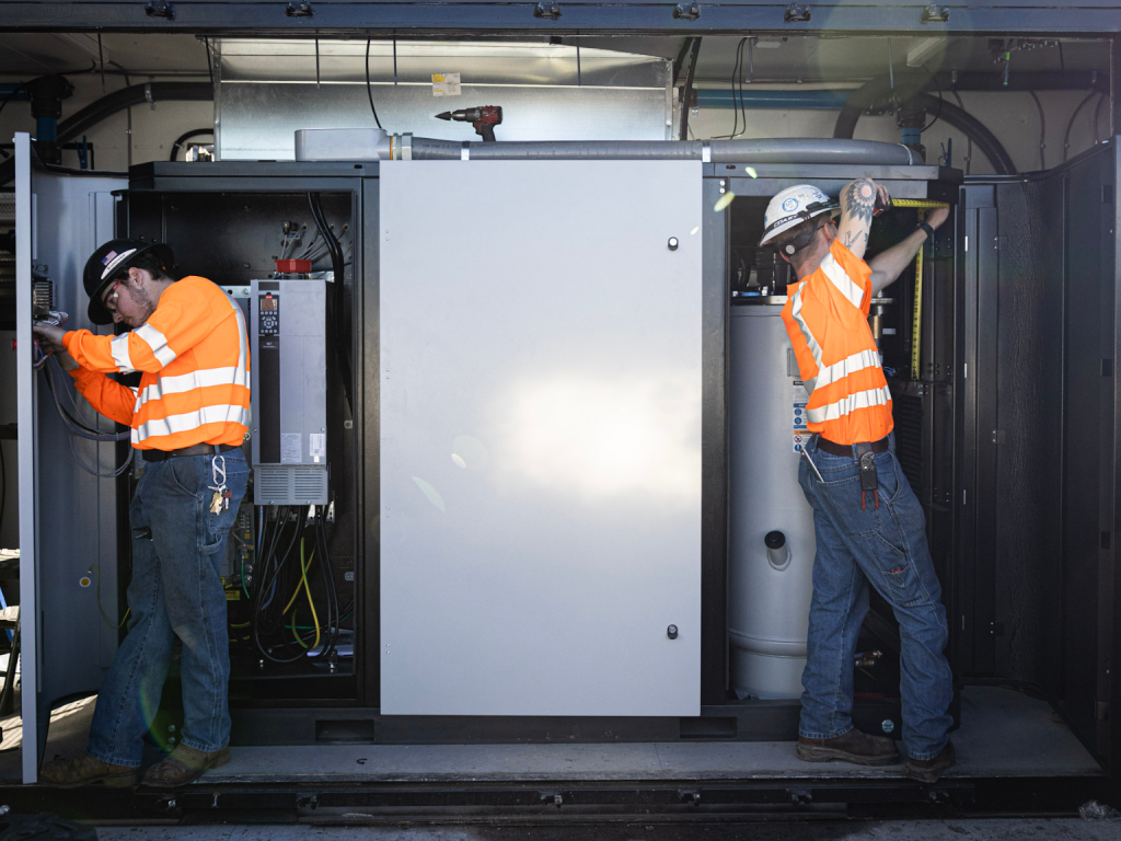 Two workers in orange safety vests and hard hats perform maintenance inside an industrial electrical cabinet enclosure.