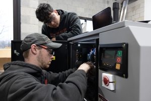 Two technicians in hoodies wearing safety glasses work on an open industrial control panel.